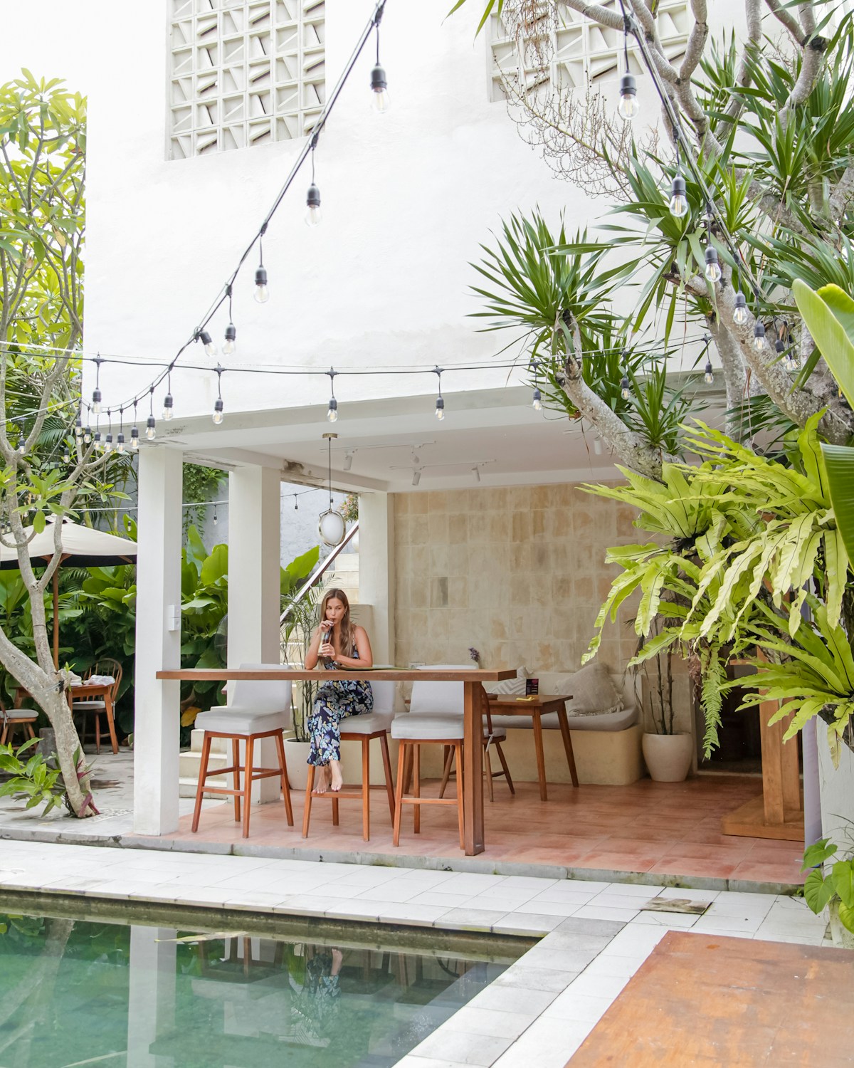 Woman working at a café table beside a pool with tropical plants, Canggu, Bali
